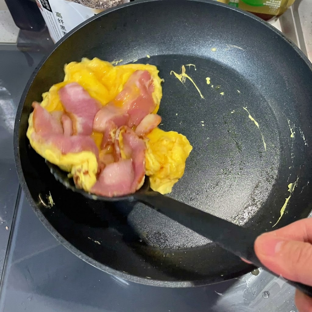 Ein Pfannenwender hebt einen gegarten Ei-Bacon-Stapel aus einer Pfanne, um ihn auf eine getoastete English-Muffin-Hälfte zu legen.