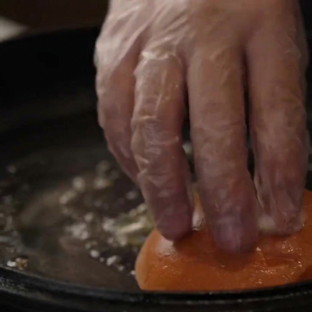 Eine be Handschuh bedeckte Hand drückt Burgerbrötchen zum Toasten in eine mit Butter benetzte Gusseisenpfanne.