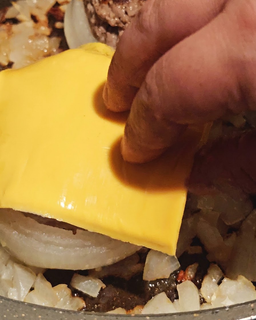 Eine Scheibe gelber Käse wird oben auf einen gegarten, fleischgefüllten Zwiebelring in der Pfanne gelegt.