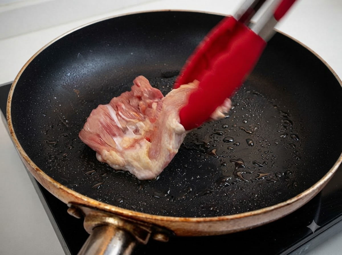 Ein roher Hähnchenschenkel wird mit roten Zangen in eine erhitzte Pfanne mit Öl gelegt.