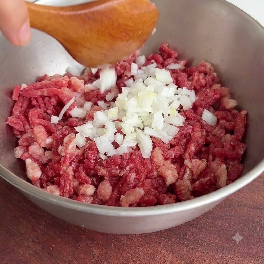 Eine Portion frisch gehackte weiße Zwiebeln wird von einem Löffel in eine Metallschüssel mit rohem Rinderhack gegeben.
