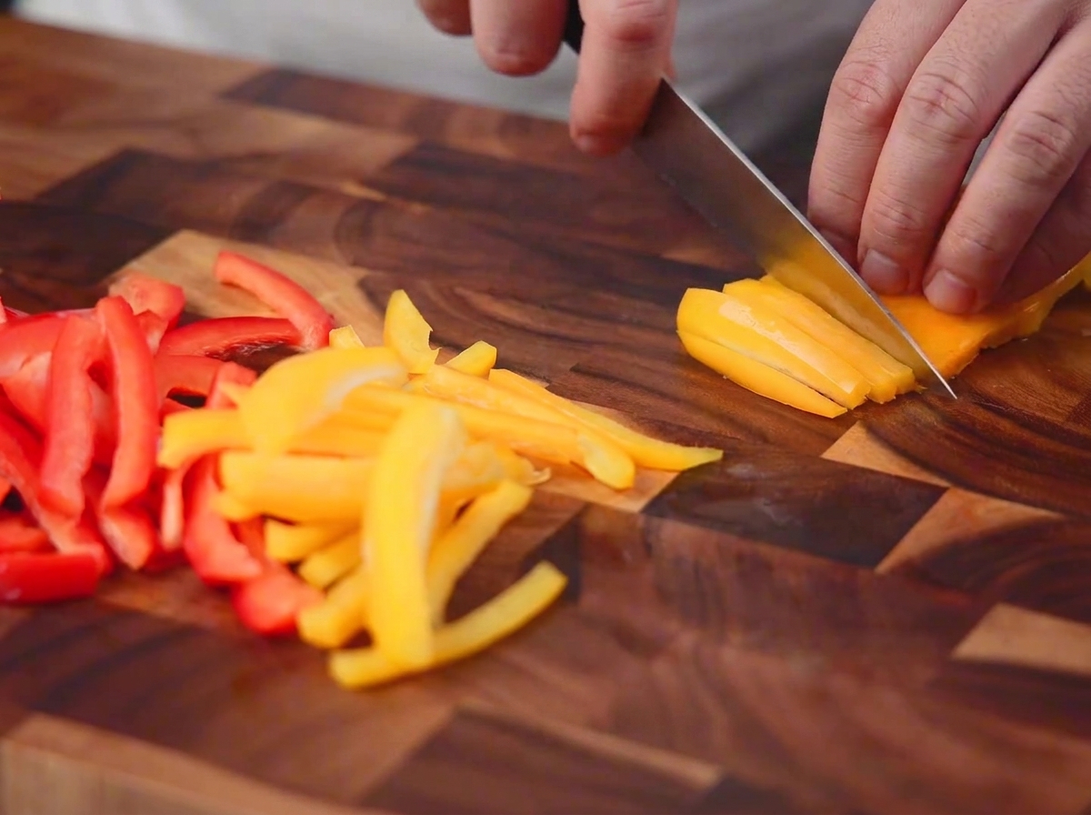 Rote und gelbe Paprika werden mit einem Messer auf einem Holzbrett in dünne Streifen geschnitten.