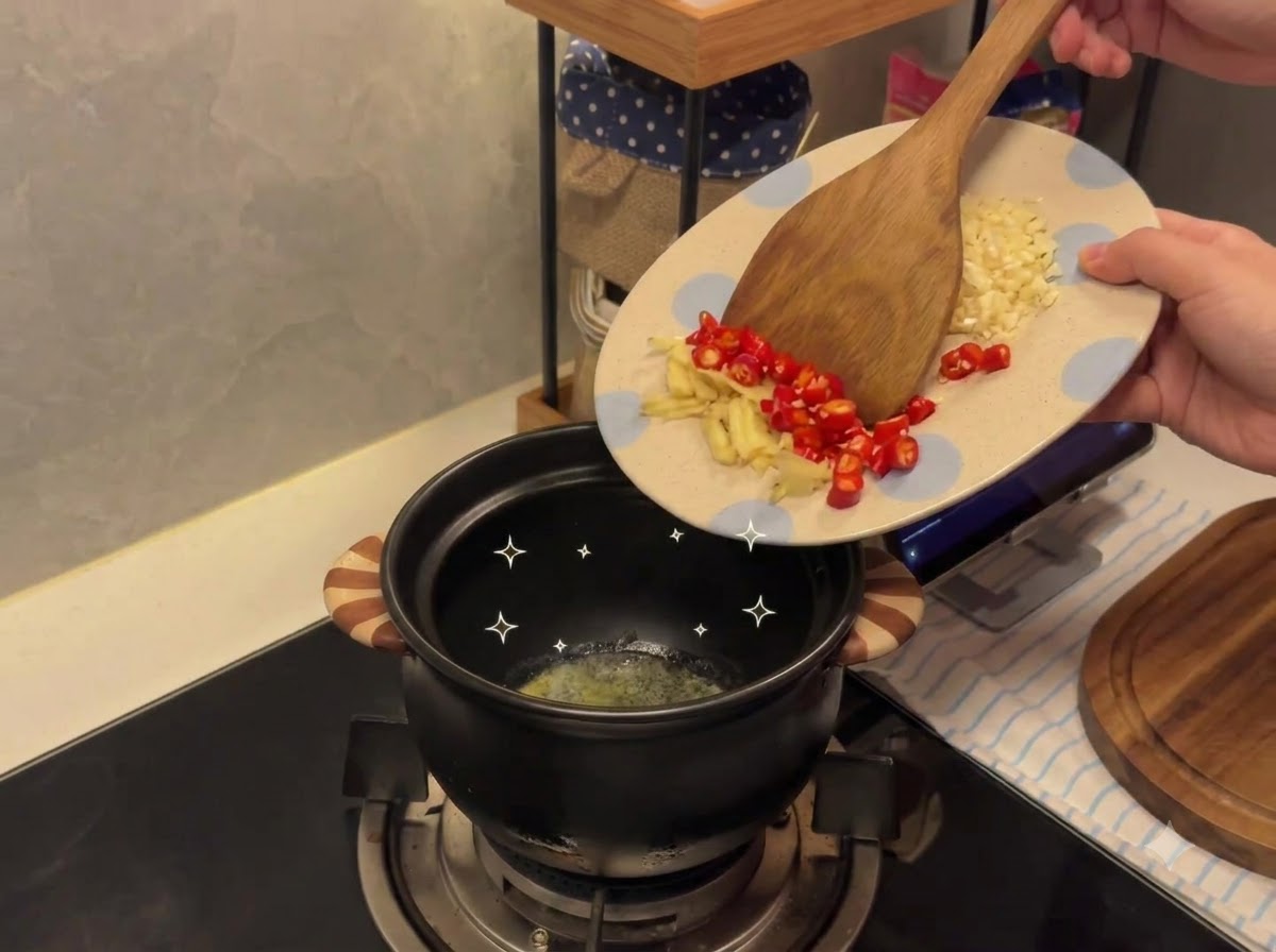Gehackter Knoblauch und Chilischoten werden von einem gemusterten Teller in einen geschmolzene Butter enthaltenden Keramiktopf gegeben.