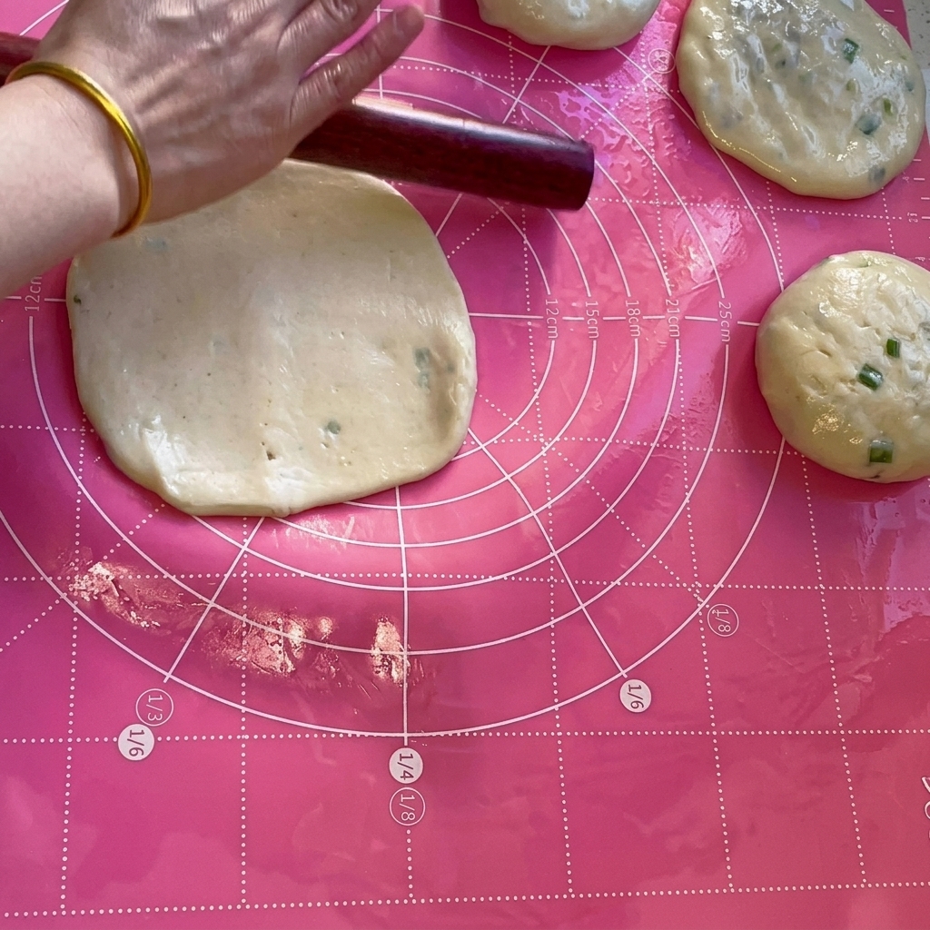 Ein hölzernes Nudelholz walzt eine Teigportion auf einer rosafarbenen Matte zu einem dünnen Kreis aus.