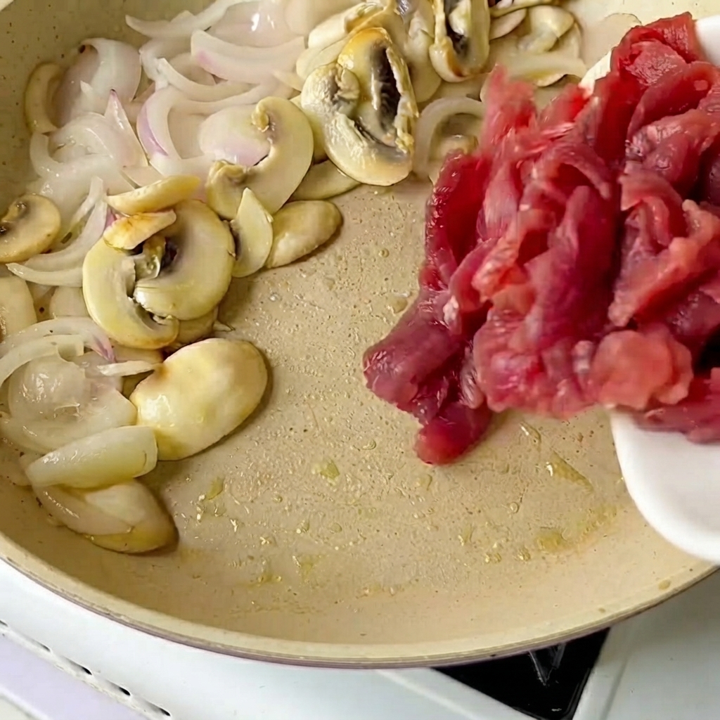 Geschnittenes Rindfleisch wird von einem weißen Teller in eine Bratpfanne mit angebratenen Zwiebeln und Pilzen gegeben.