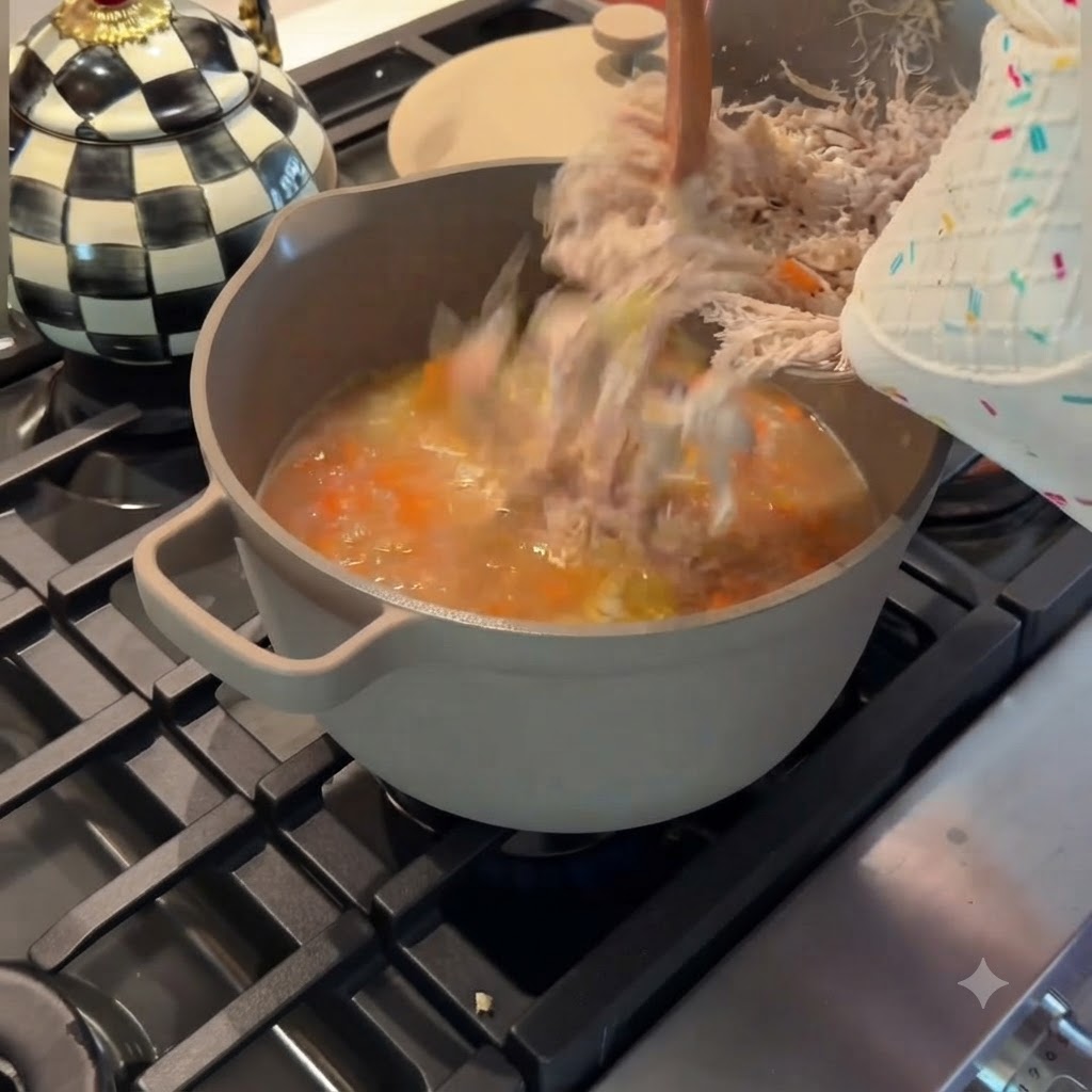 Fein zerzupftes Hähnchen, das aus einer Metallschüssel zurück in einen grauen Topf mit köchelnder Hühner-Gemüse-Suppe gegossen wird.