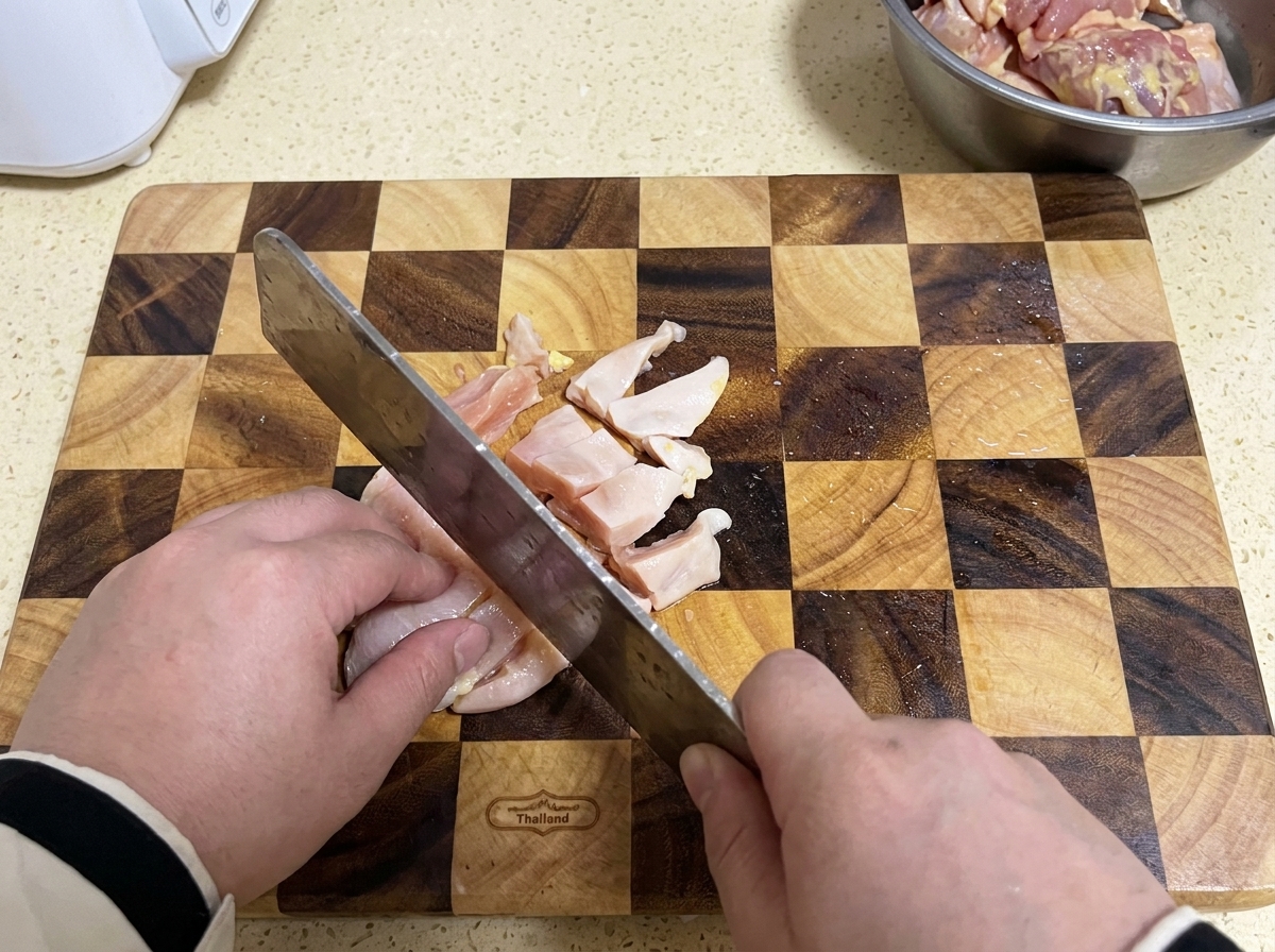 Ein Beil schneidet rohe Hühnerbrust in kleine Stücke auf einem karierten Holzschneidebrett.