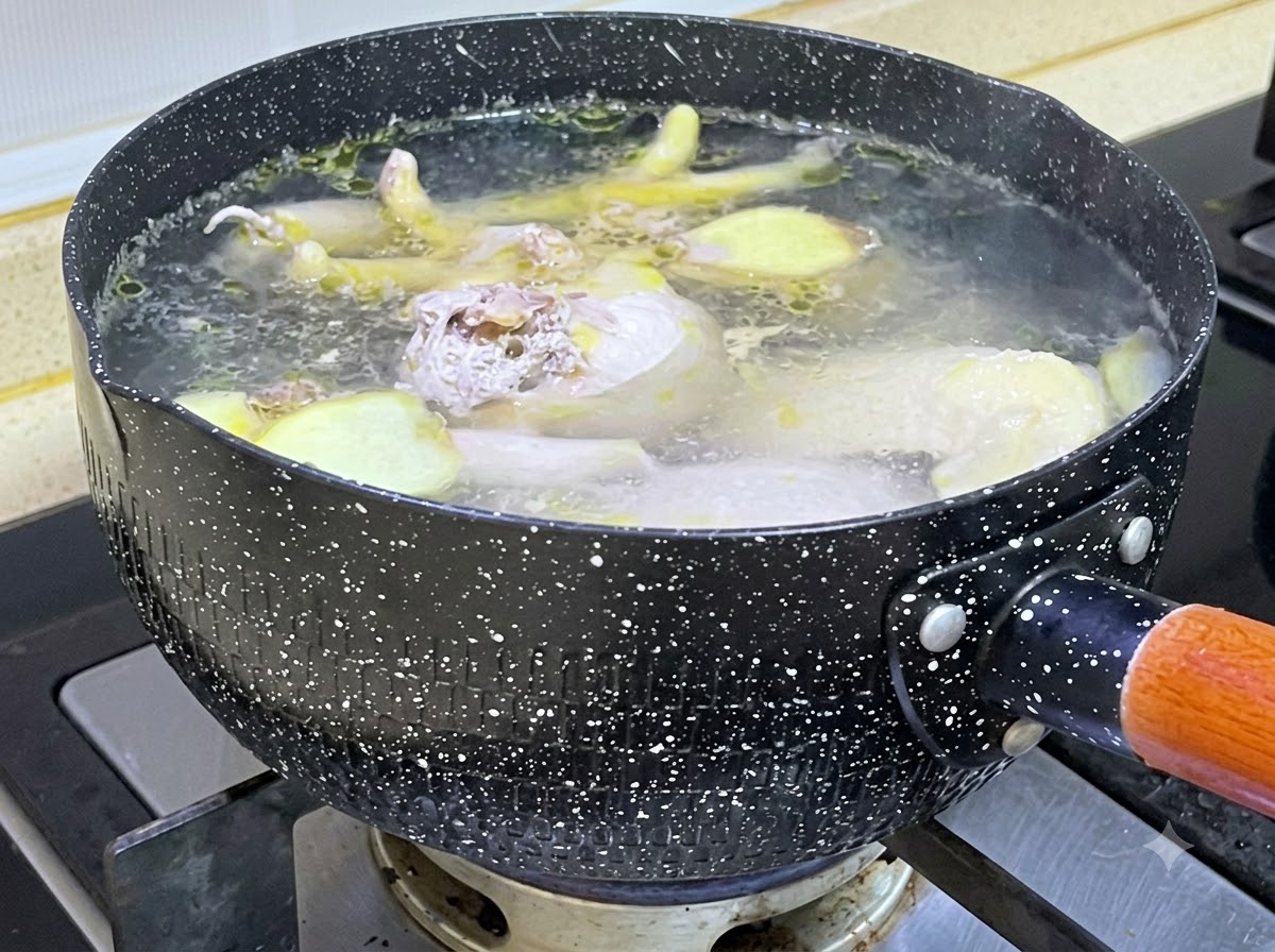 Rohe Hühnerstücke und Ingwerscheiben köcheln in einem schwarz gesprenkelten Topf mit kochendem Wasser.