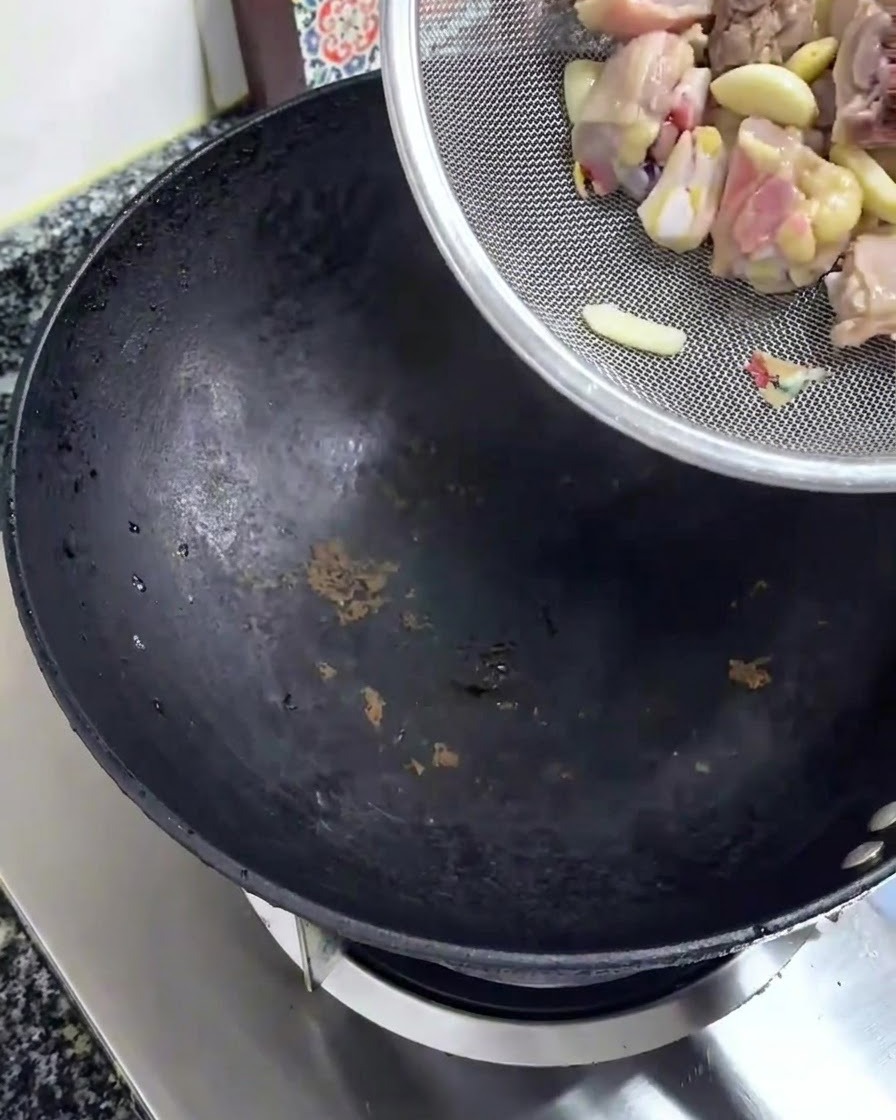 Rohe gehackte Hühnerstücke werden aus einem silbernen Sieb in einen schwarzen Wok geschüttet.