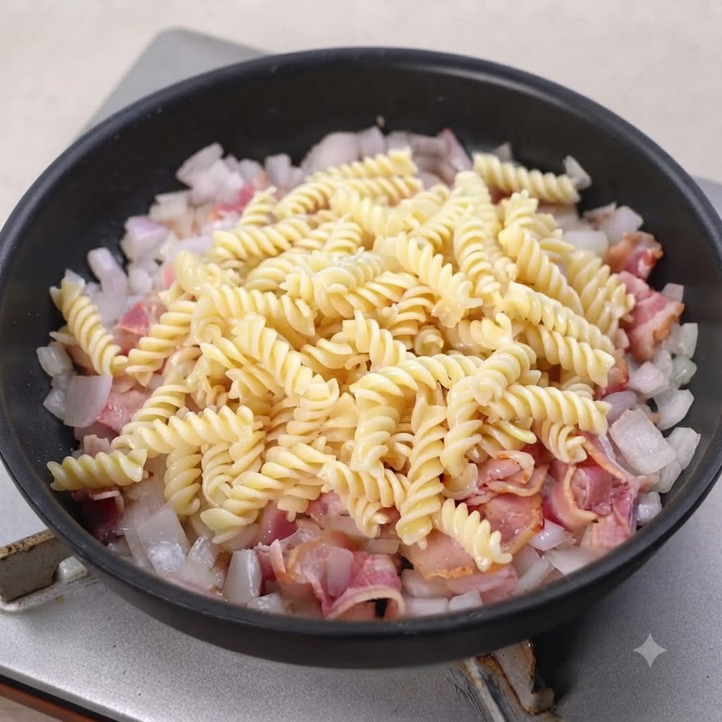 Gekochte Spiralnudeln liegen auf einer Schicht aus gebratenem Speck und Zwiebeln in einer schwarzen Pfanne.
