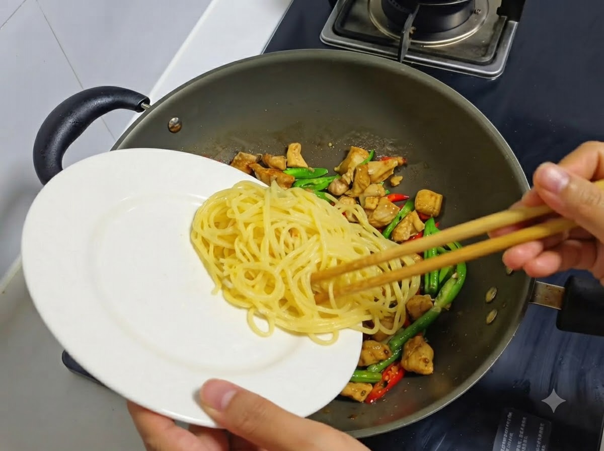 Gekochte Spaghetti-Nudeln werden von einem weißen Teller in einen Wok mit gebratenem Hähnchen und Chilis gegeben.