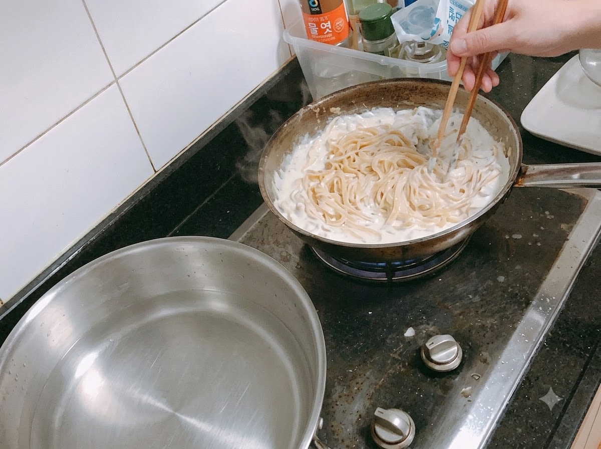 Pasta wird mit Essstäbchen in einer Pfanne unter eine köchelnde Sahnesauce gemischt.