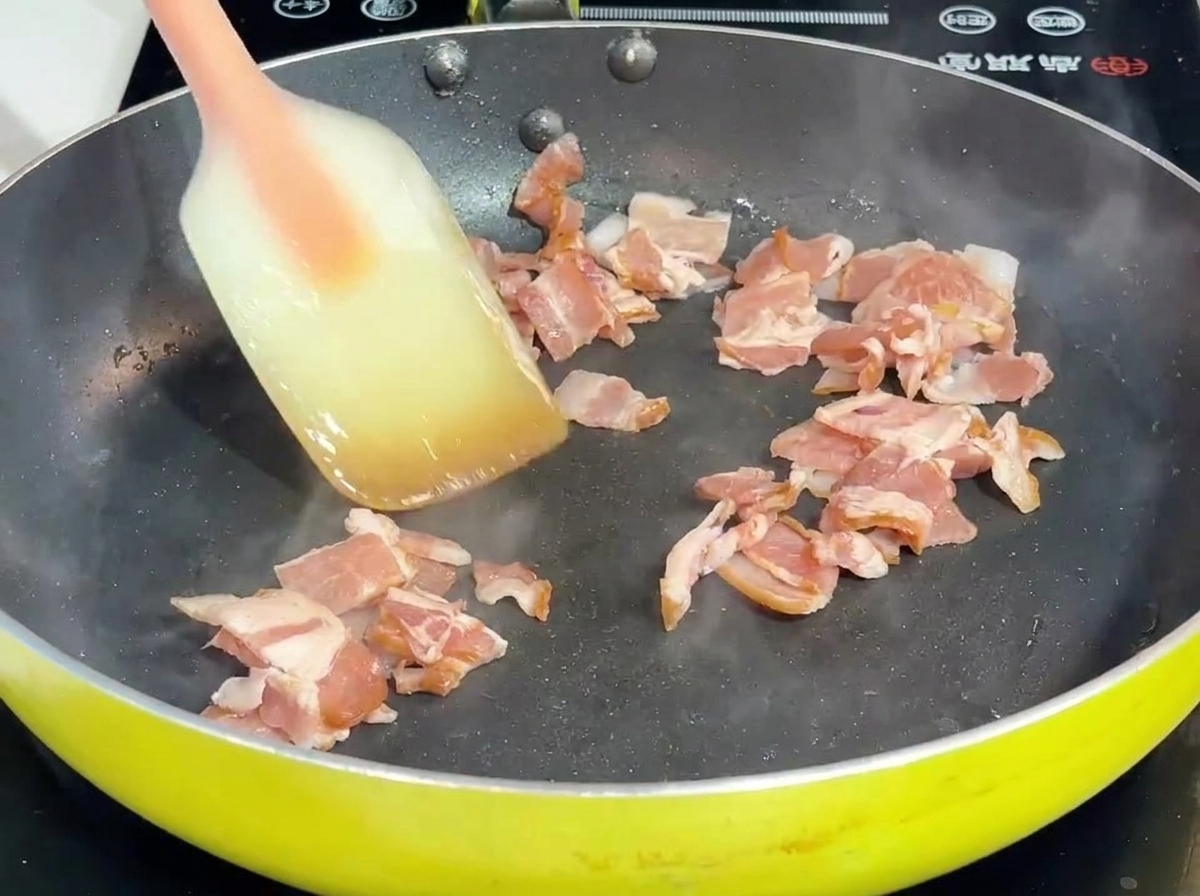 Speck und Zwiebel werden mit einem Spatel in einer Pfanne angebraten.