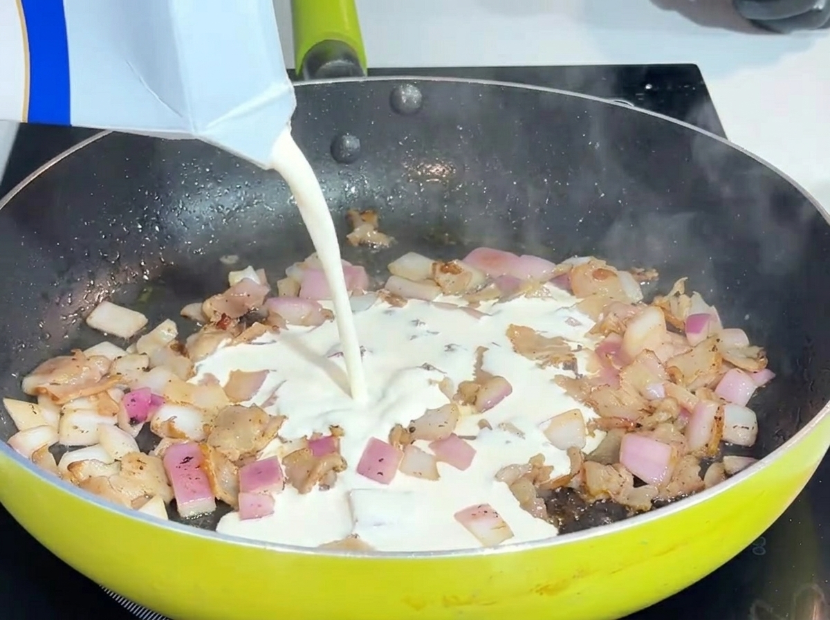 Flüssige Schlagsahne wird in eine Pfanne mit Speck und Zwiebeln gegossen.