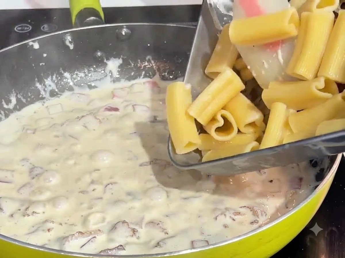 Vorgekochte herzförmige Pasta wird aus einer Schale in eine blubbernde Creme-Speck-Sauce in einer Pfanne gegeben.