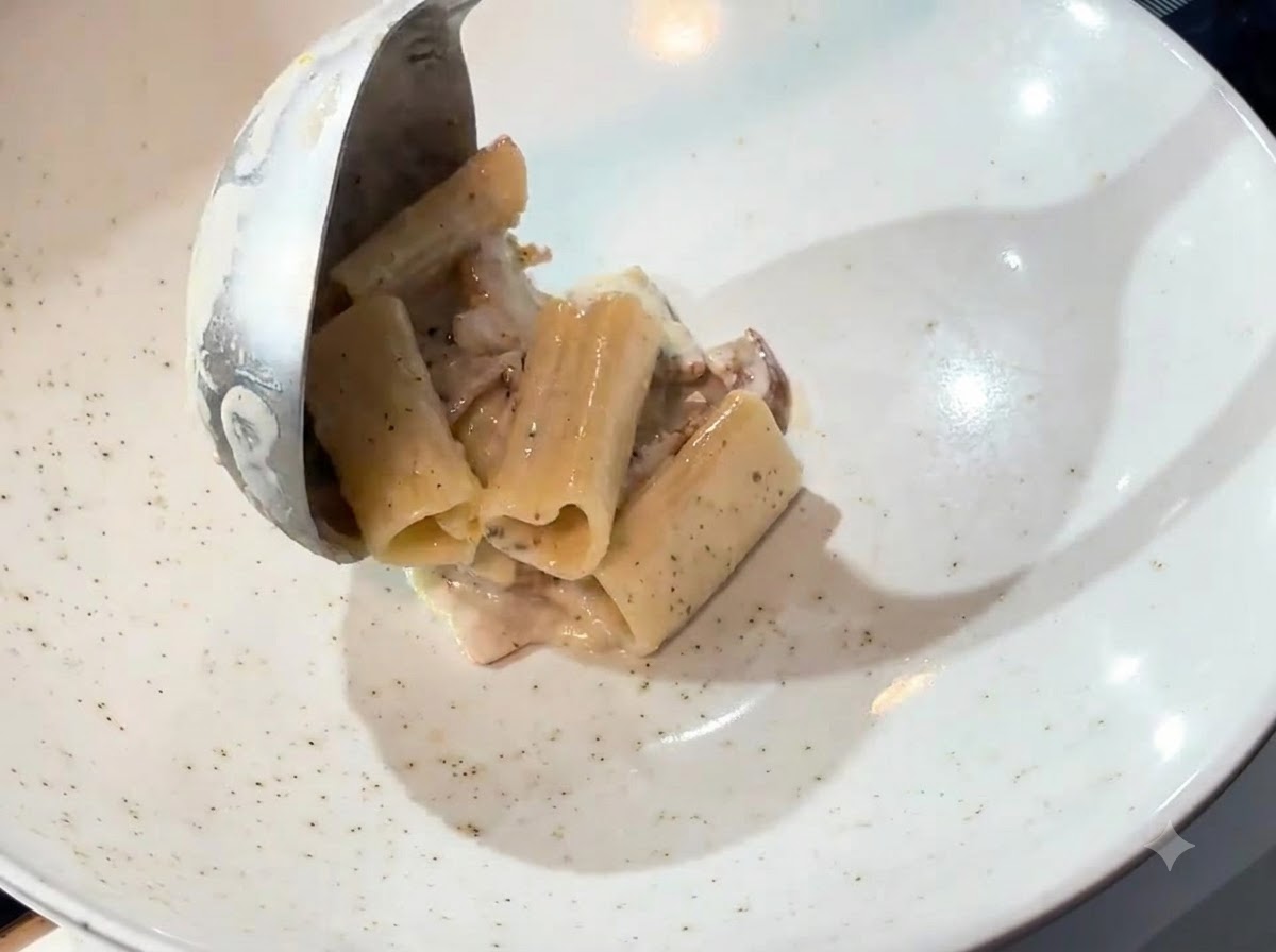 Eine Schöpfkelle transportiert cremige Pasta aus einer Pfanne in eine weiße Servierschüssel.