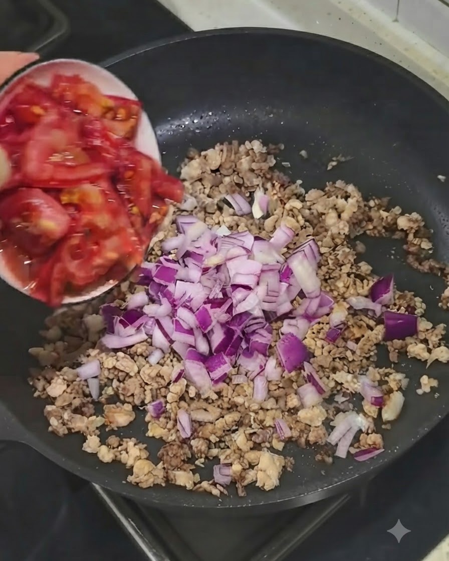 Frische gewürfelte Tomaten werden aus einer kleinen Schüssel in die Pfanne mit Fleisch und Zwiebeln gegeben.