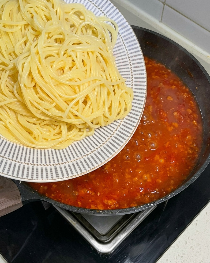 Frisch gekochte Spaghetti werden von einem gemusterten Teller in eine große Pfanne mit blubbernder roter Fleischsoße überführt.