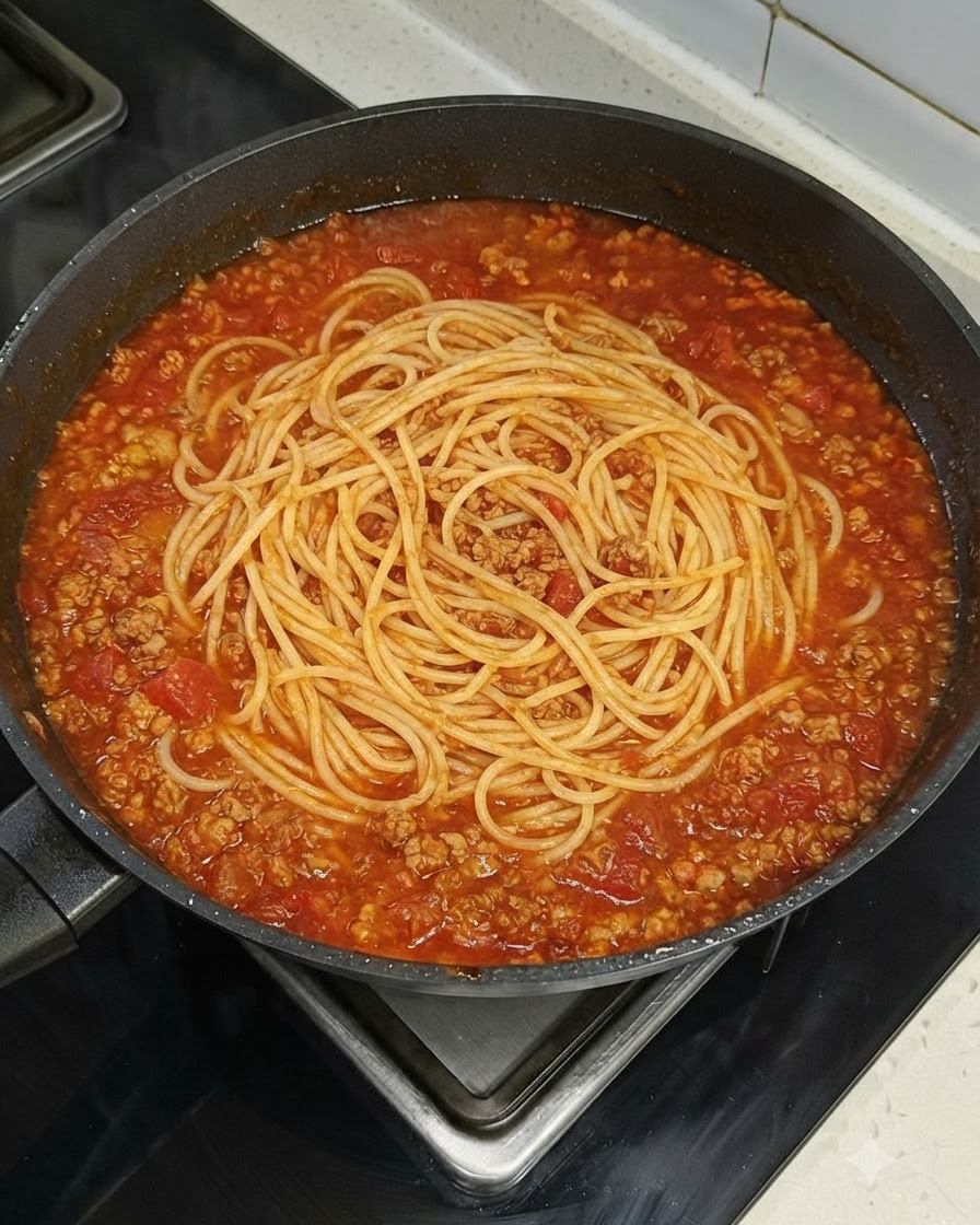 Ein Nest aus Spaghetti-Nudeln liegt gleichmäßig in einer großen Pfanne mit roter Fleischsoße auf einem Herd.