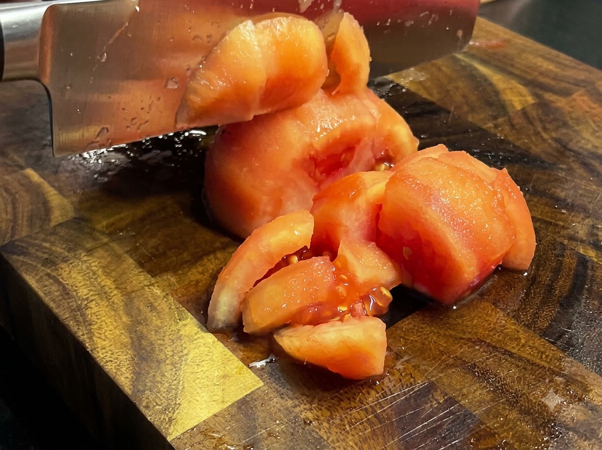 Ein Küchenmesser hackt gekochte, geschälte Tomaten in Stücke auf einem Holzschneidebrett.