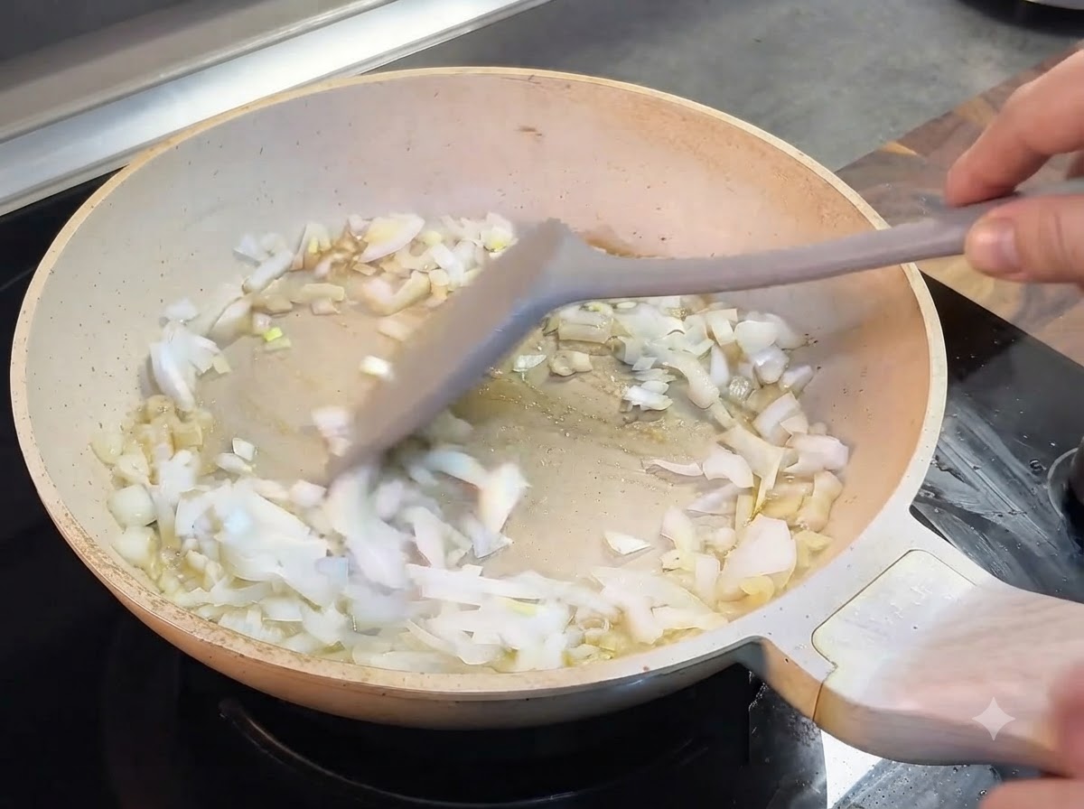Gehackte Zwiebeln werden mit einem grauen Spatel in einer hellen Pfanne umgerührt.