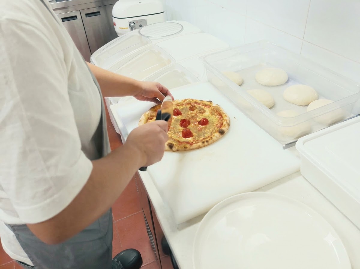 Eine Person benutzt einen Rollschneider, um eine frisch gebackene Pizza auf einem weißen Schneidebrett zu schneiden.