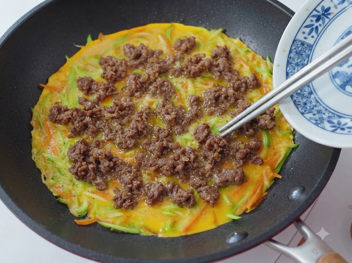 Nahaufnahme von Stäbchen, die gekochte Rindfleischkrümel auf einer Pfanne mit Gemüse- und Eipfannkuchenboden verteilen.