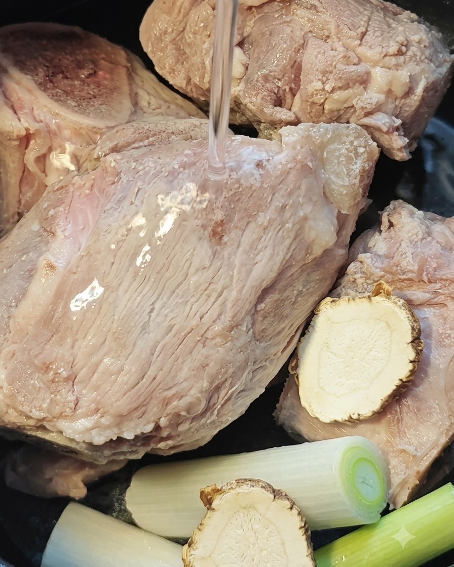 Wasser wird über Schweineknochen, Angelikawurzel und Lauchzwiebeln in einem Reiskochertopf gegossen.