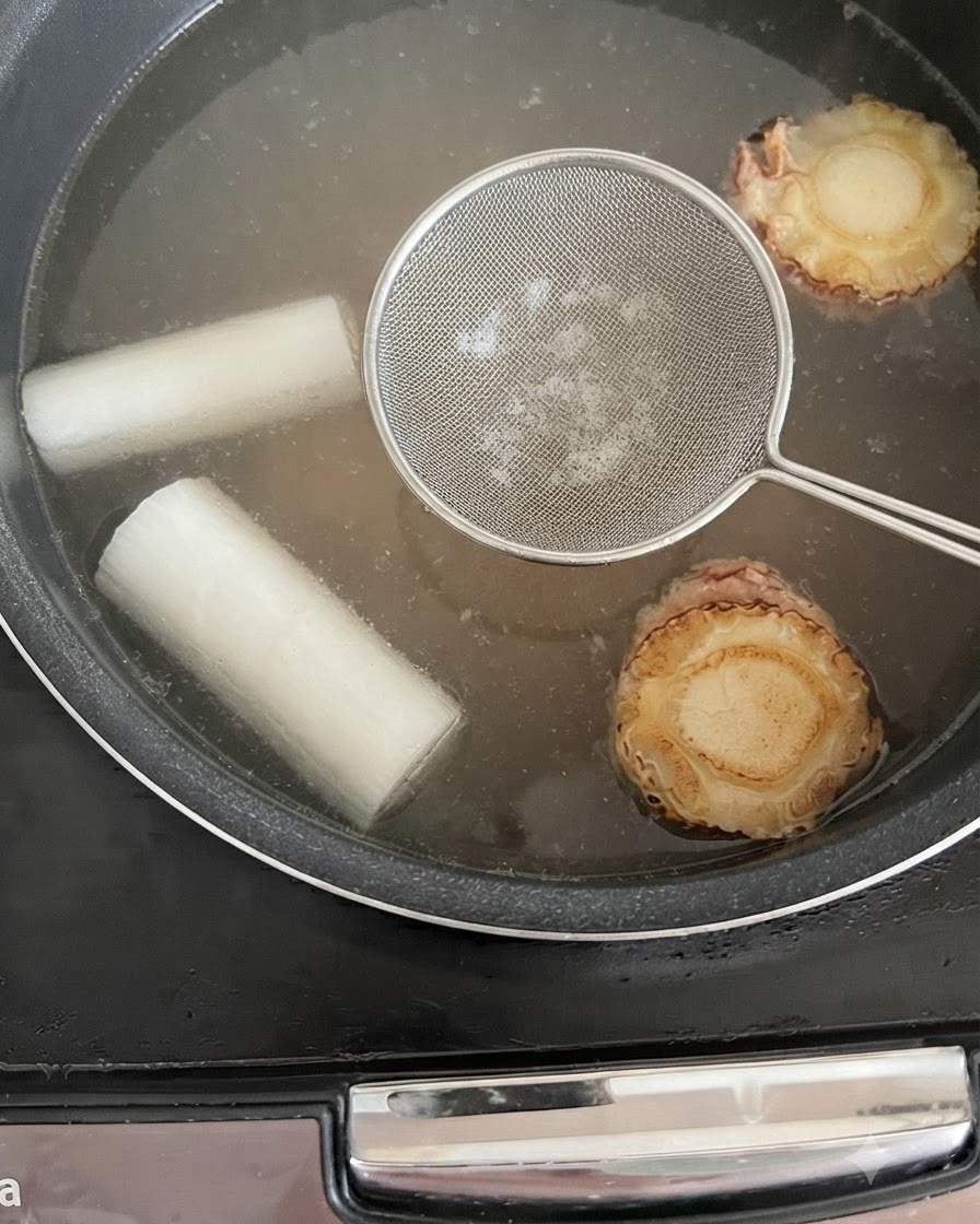 Ein Schaumlöffel über der köchelnden Brühe im Reiskocher, um Unreinheiten zu entfernen.