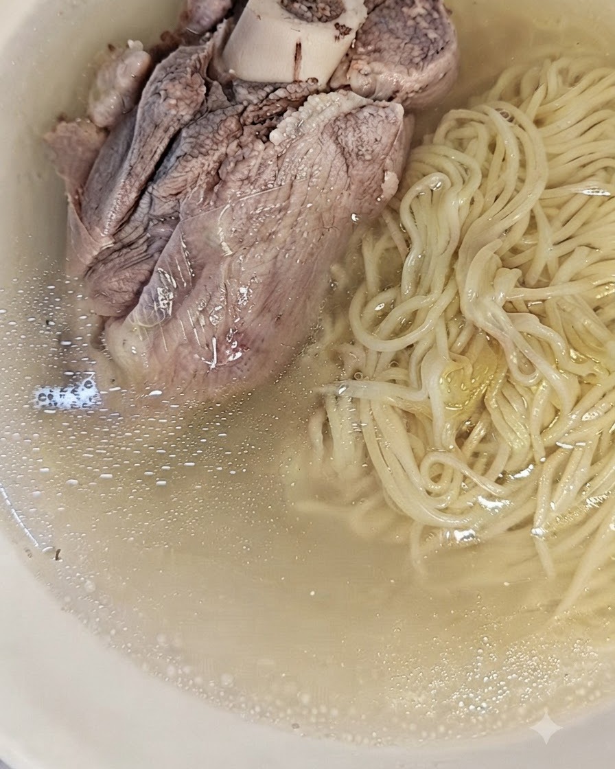 Gekochte Nudeln in einer Schüssel neben der klaren, abgesiebten Schweinebrühe.