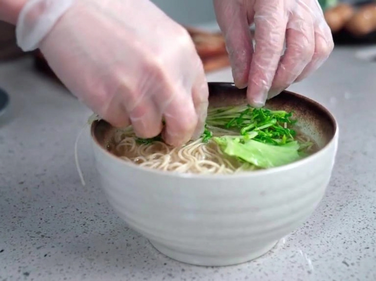 Behandschuhte Hände legen vorsichtig frisches, grünes Blattgemüse in eine dampfende Schüssel mit Ramen-Nudeln und milchiger Brühe.