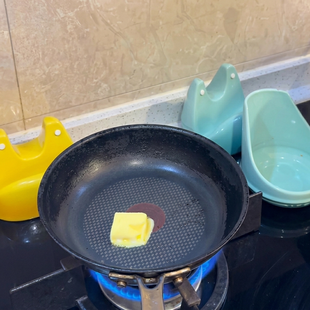 Ein gelber Butterwürfel schmilzt in der Mitte einer schwarzen Antihaft-Pfanne über einer Gasflamme.