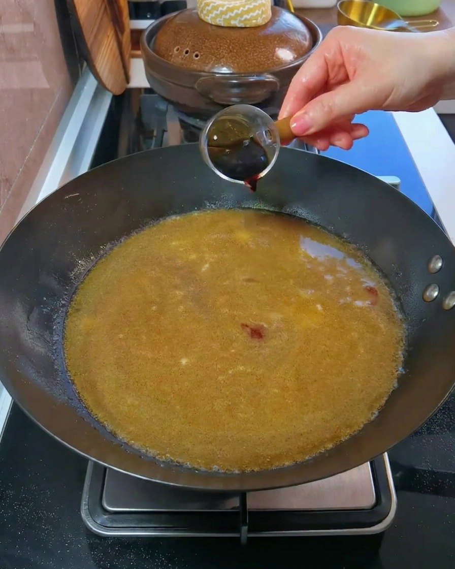 Eine Hand gießt dunkle Sojasoße aus einem kleinen Messbecher in einen mit Brühe gefüllten Wok.