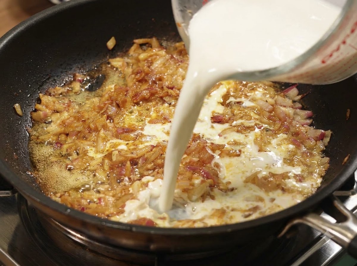 Milch wird aus einem Messbecher in eine Pfanne mit köchelnden Zwiebeln und Curry gegossen.