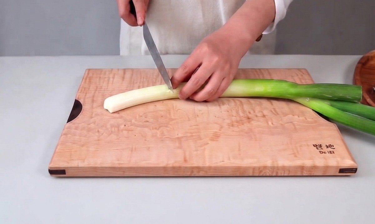 Hände halten ein Küchenmesser und schneiden eine große Frühlingszwiebel auf einem Holzschneidebrett.