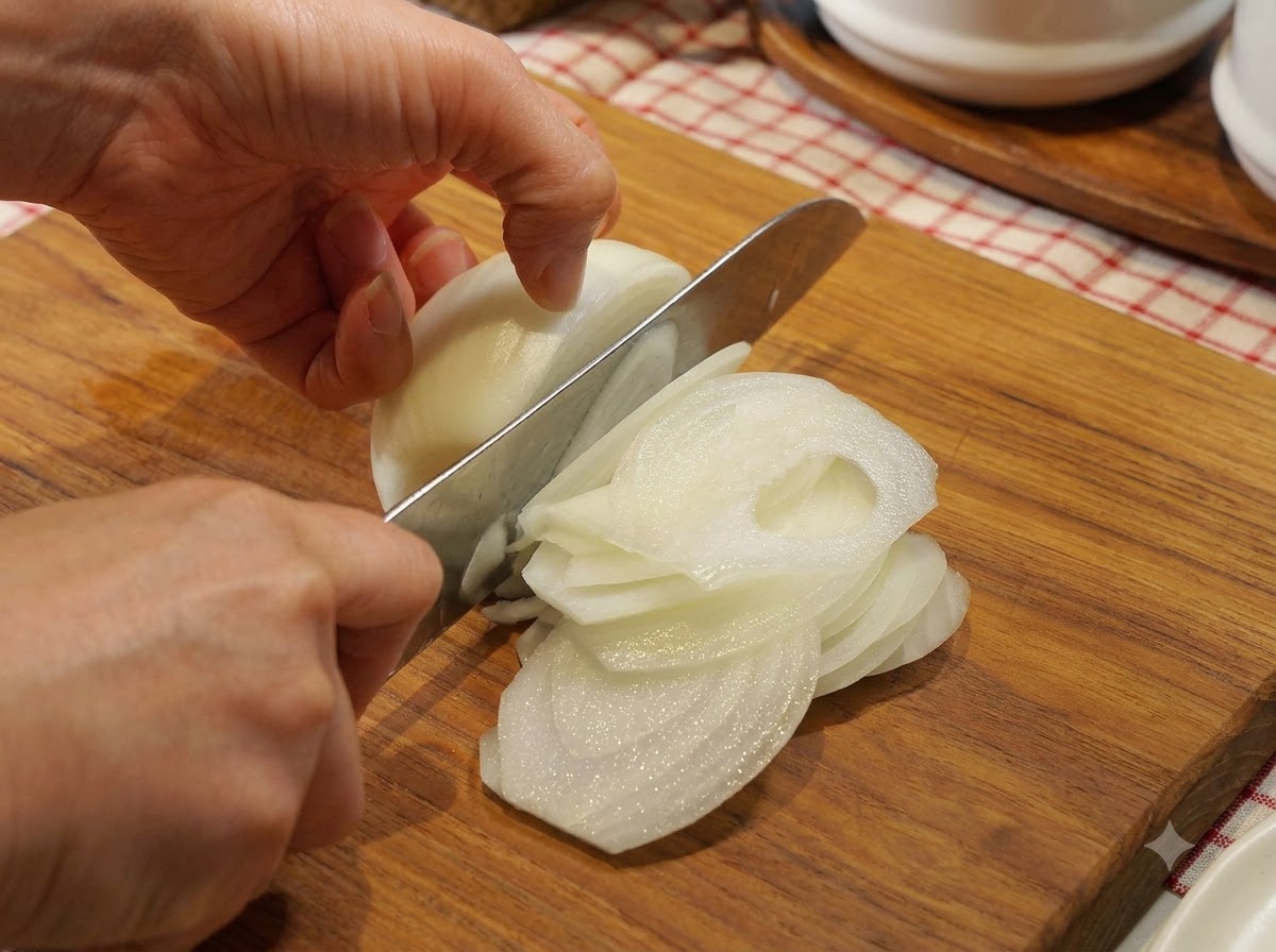 Eine Zwiebel wird auf einem Holzschneidebrett mit einem Kochmesser sorgfältig in dünne Halbmonde geschnitten.