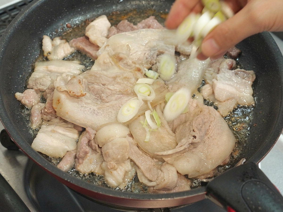 Gehackte Lauchzwiebeln werden in eine Pfanne mit bratenden Schweinebauchscheiben gegeben.