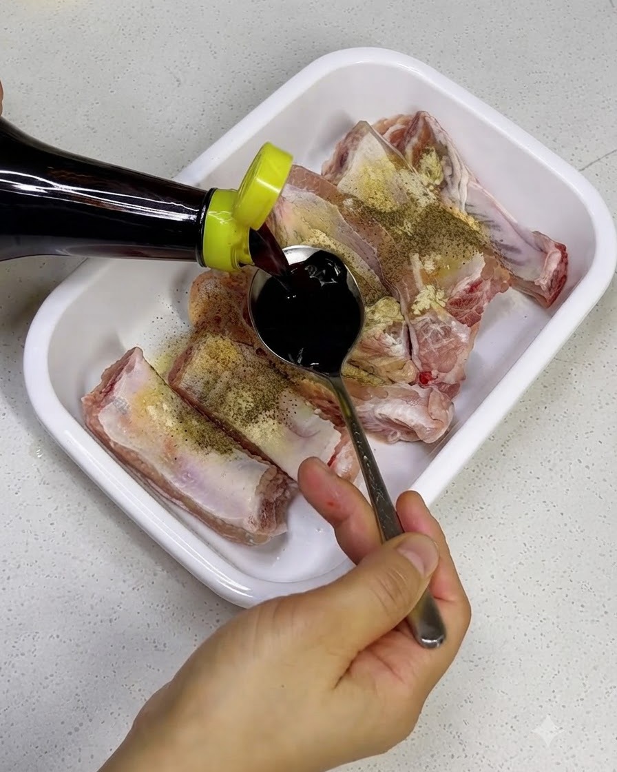 Dunkle Sojasauce wird aus einer Flasche auf rohe Schweinerippchen in einer weißen Schale gegossen.
