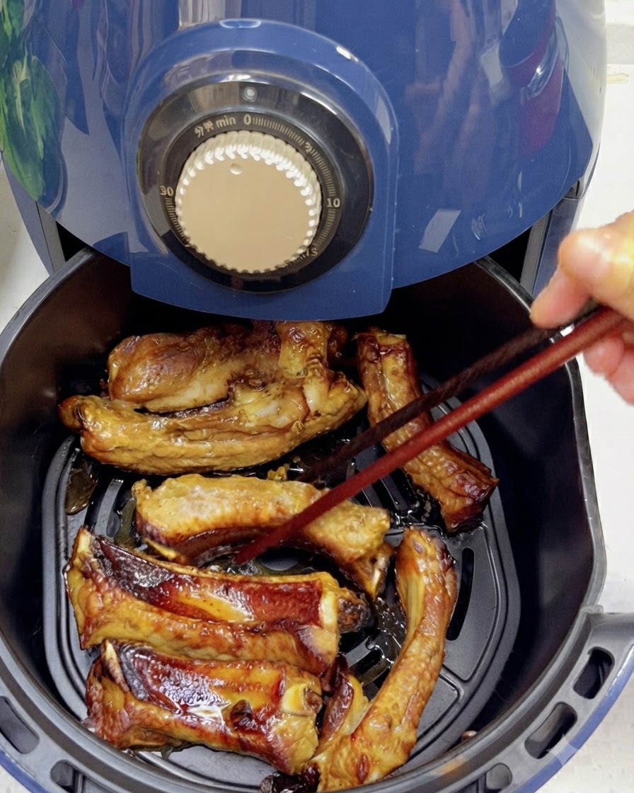 Mit Stäbchen werden teilweise gegarte, gebräunte Schweinerippchen in einem Heißluftfritteusenkorb gewendet.