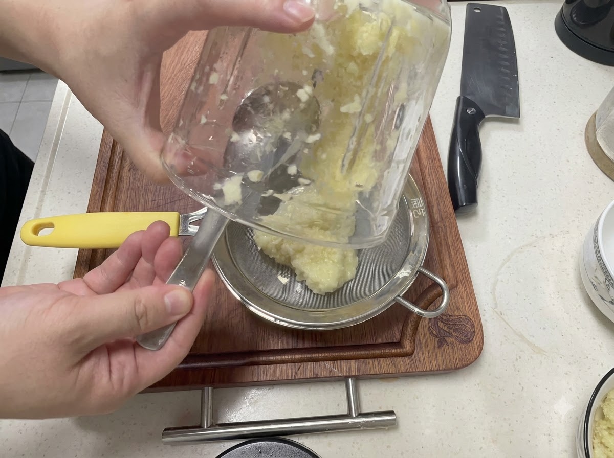 Pürierter Knoblauch wird aus einem Glasbehälter durch ein feines Sieb über eine Metallschüssel gegossen.