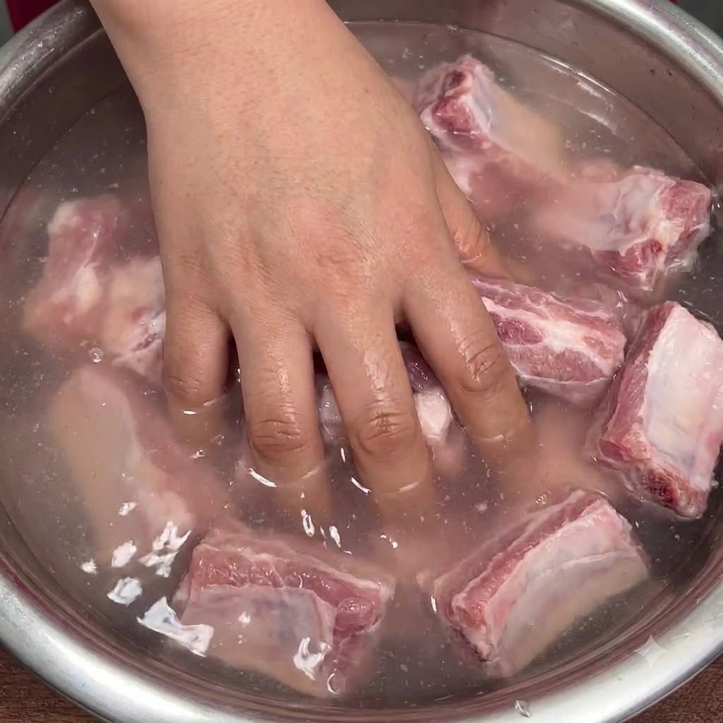 Eine Hand mischt rohe Schweinerippchen in einer Schüssel mit trübem Wasser kräftig durch, um das Natron zu verteilen.
