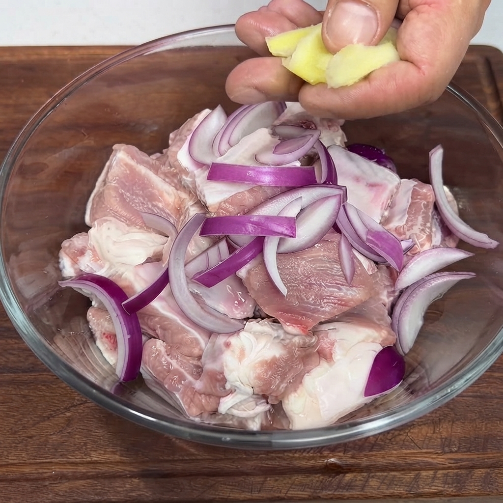 Eine Person legt Ingwerscheiben in eine Glasschüssel gefüllt mit gehackten rohen Schweinerippchen und roten Zwiebelscheiben.