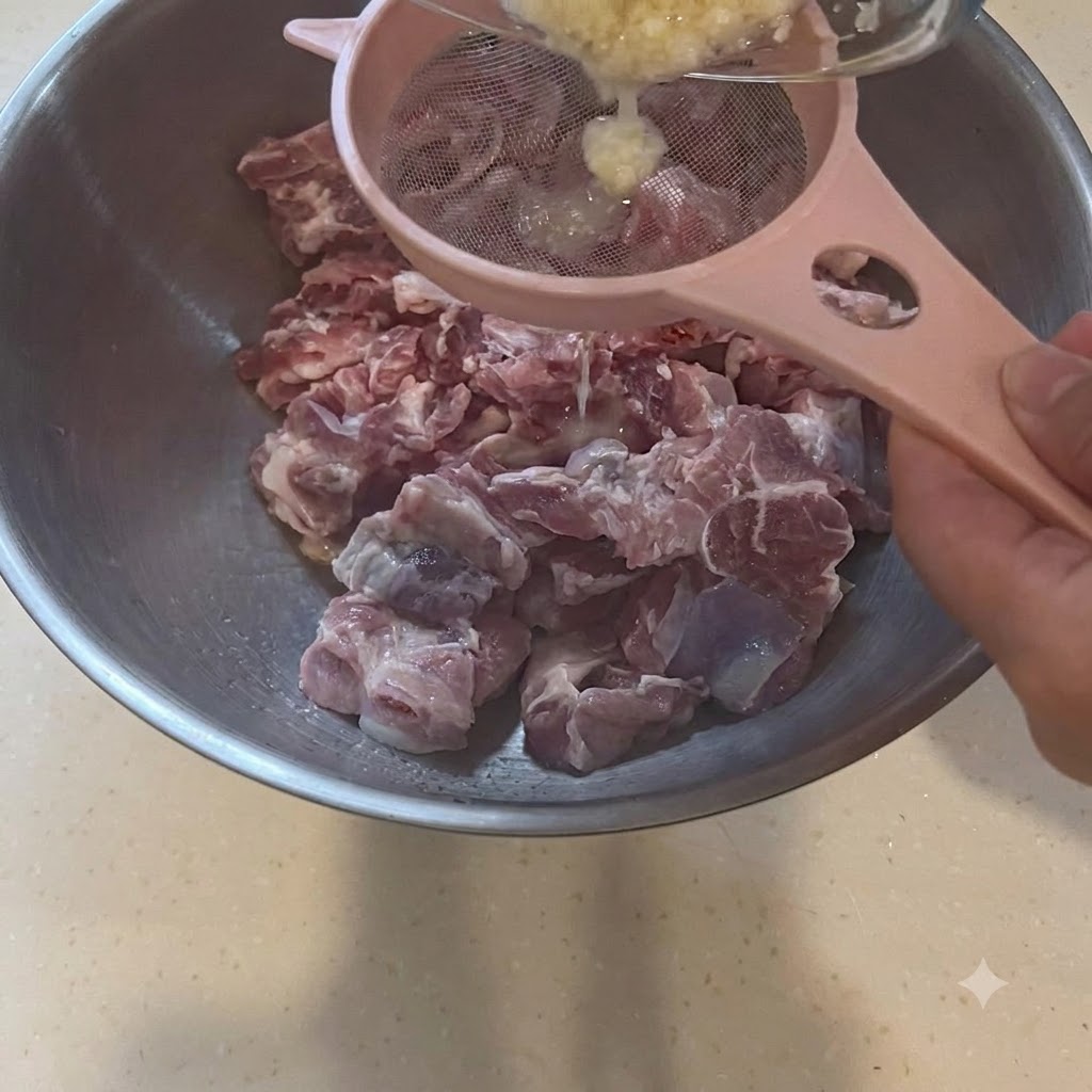 Knoblauchpaste wird durch ein rosafarbenes Sieb direkt über eine große Schüssel mit rohen Schweinestücken gegossen.