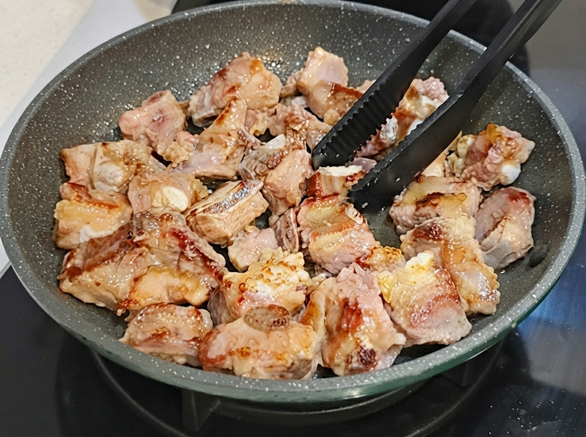 Schweinerippchen werden mit schwarzen Zangen in einer beschichteten Pfanne gewendet.