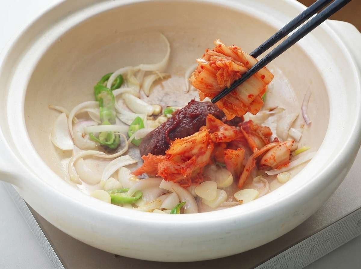 Koreanische Chilipaste und Kimchi werden mit Stäbchen in den Topf mit den angebratenen Aromaten gegeben.