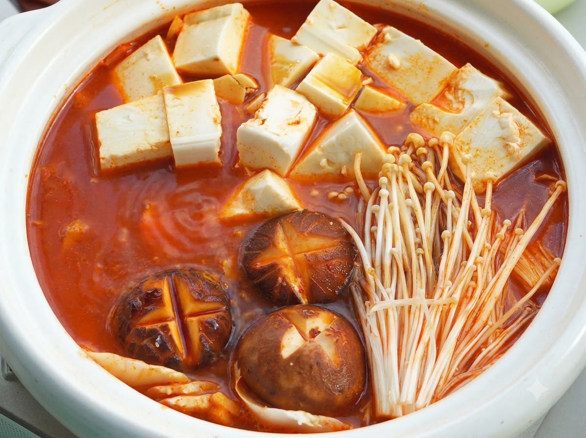 Ein weißer Keramikhaltpott gefüllt mit köchelnder roter Suppenbasis, weichen Tofuwürfeln, Shiitake-Pilzen mit Einschnitten und Enoki-Pilzen unter einem Deckel.