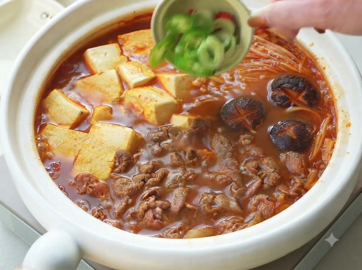 Frühlingszwiebeln und geschnittene Chilischoten werden aus einer kleinen Schüssel als Garnitur über den gekochten koreanischen Rinder- und Tofueintopf gestreut.