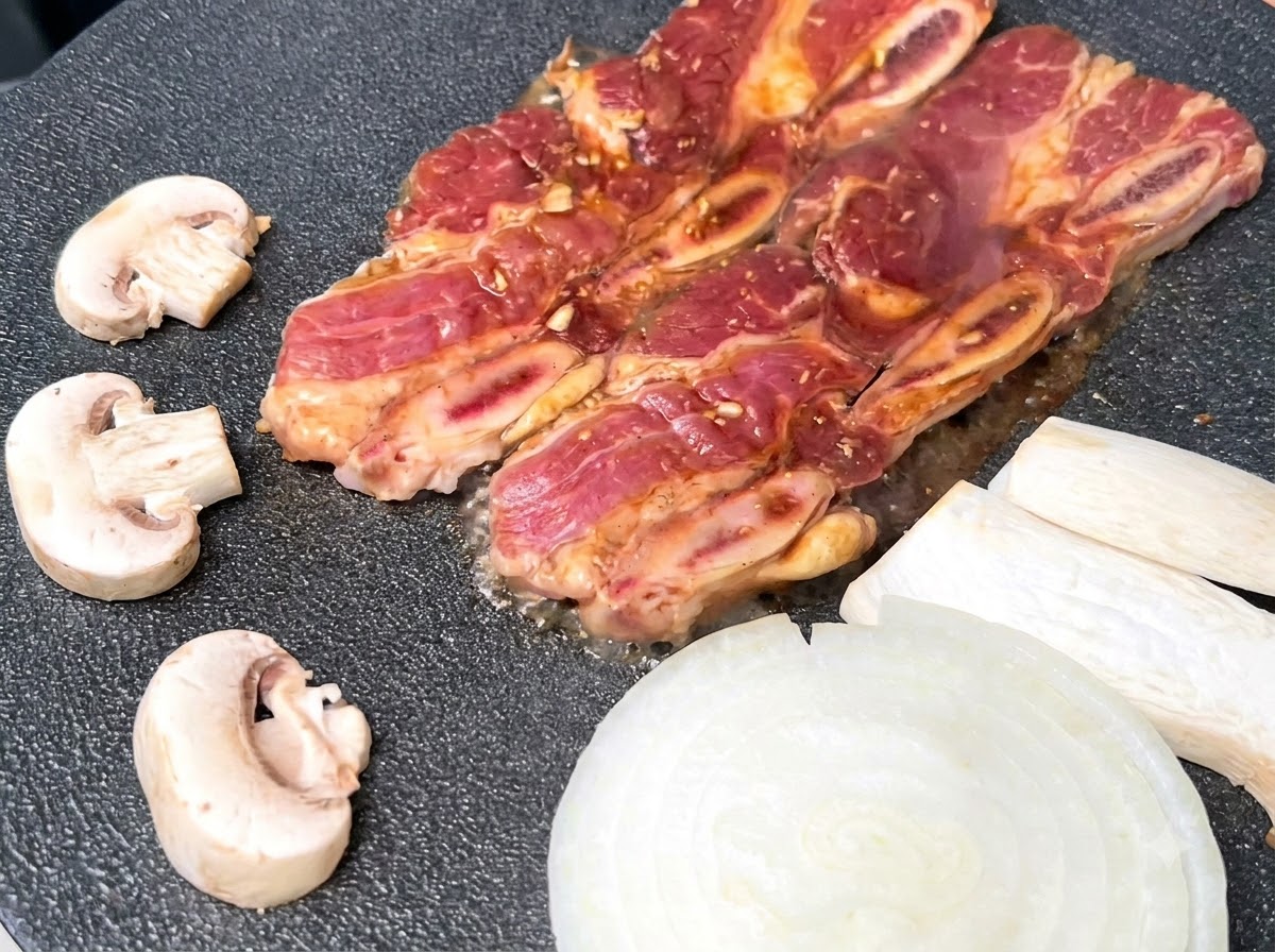 Brutzelnde Rinderrippchen auf einer heißen schwarzen Grillpfanne neben Zwiebeln und Pilzen.