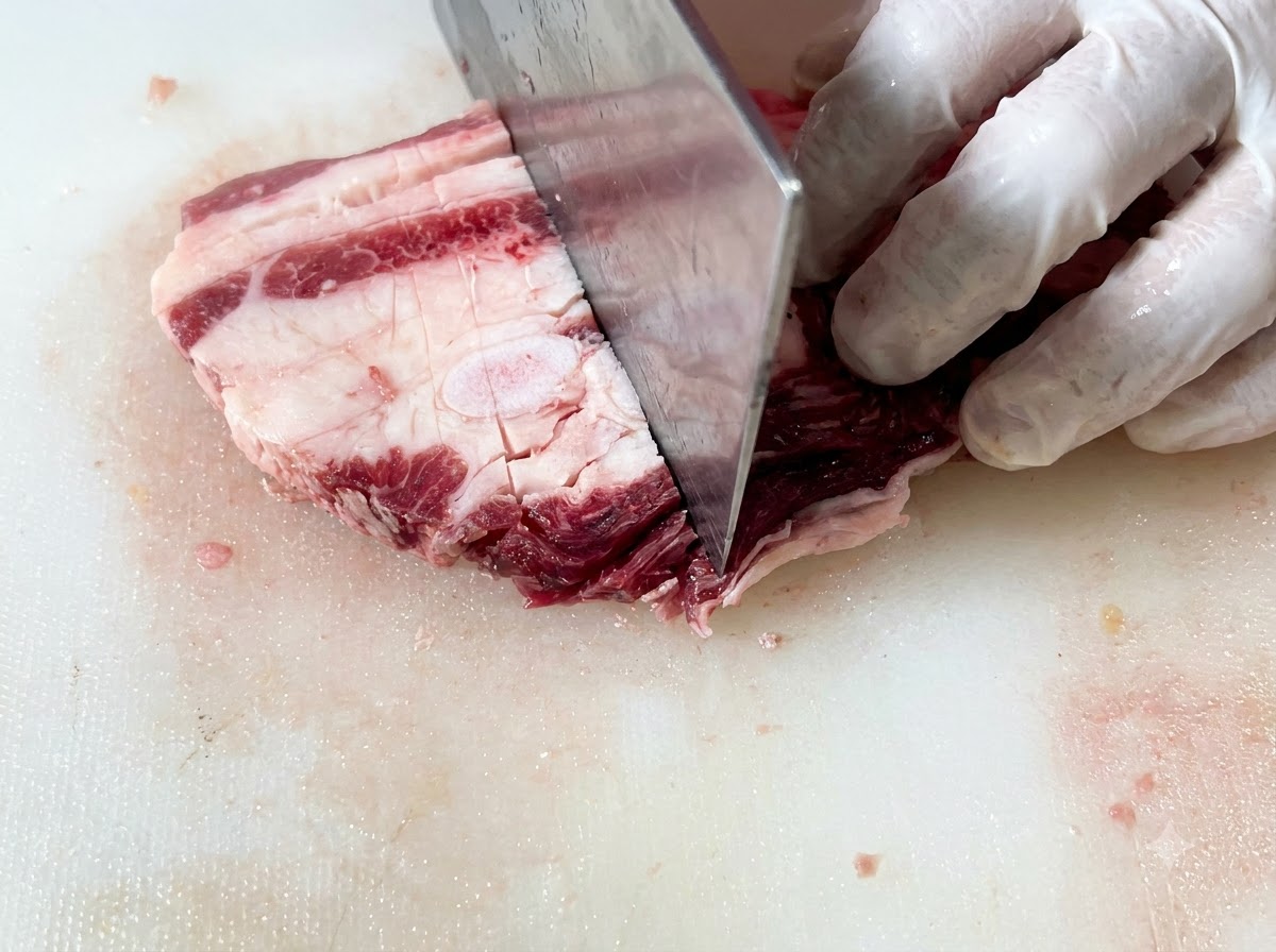 Rohe Rinder-Short-Ribs werden auf einem Schneidebrett mit einem großen Messer in gleichmäßige rechteckige Stücke geschnitten.