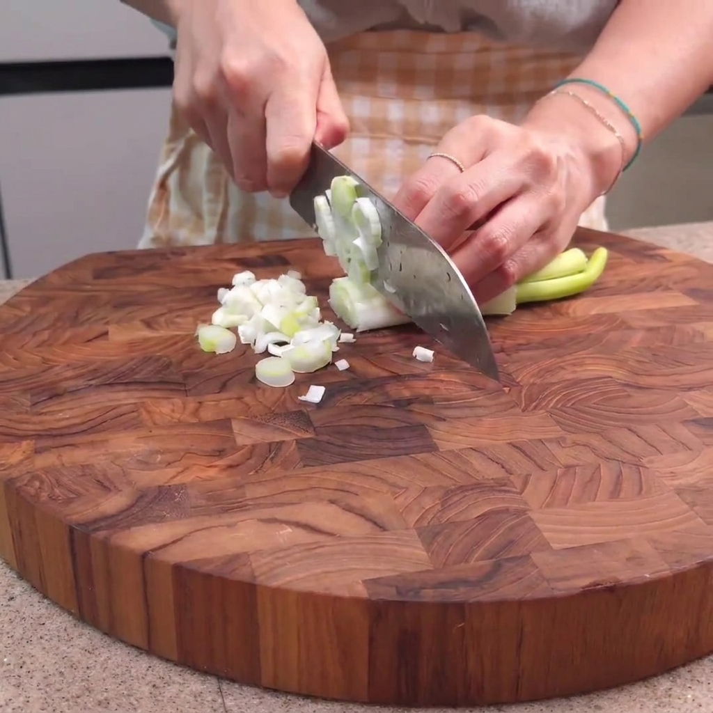 Frühlingszwiebeln werden auf einem Holzschneidebrett mit einem Kochmesser zerkleinert.